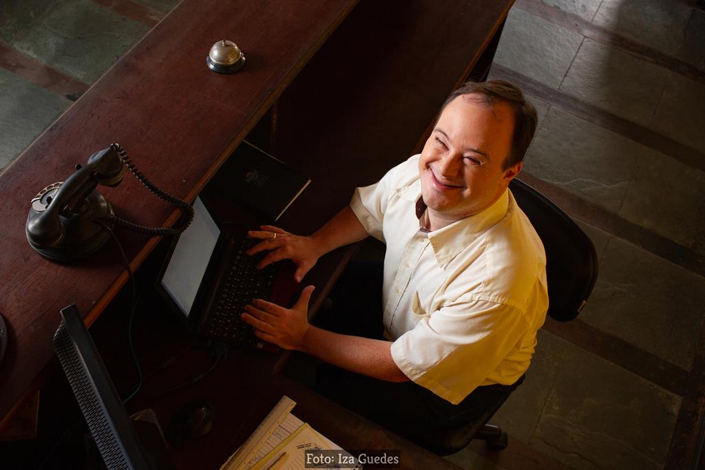 Foto de homem branco com sindrome de down utilizando notebook em mesa e sorri para câmera